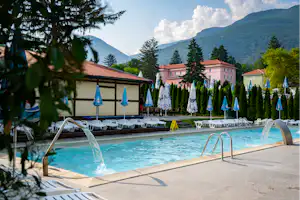 Schwimmbad mit Wassersprudlern und Liegen am Rand, dahinter ein Gebäude und Berge in der Ferne.