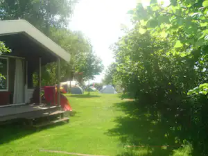 Wohnwagen auf einem Campingplatz mit Terrasse und Rasenfläche, im Hintergrund ein weiterer Zeltplatz zwischen Bäumen.