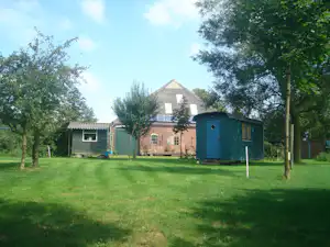 Campingplatz mit Rasenweg, Backsteinhaus und Garten, daneben ein Schuppen und grüne Bäume im Hintergrund.