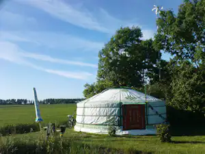 Zelt auf einem Campingplatz am Rand eines Feldes, mit Holzschrank und Campingstühlen, umgeben von Bäumen und blauem Himmel.