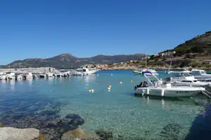 Hafen mit vielen Booten im klaren Wasser, Steg und Felsen, im Hintergrund Berge und Häuser am Hang.