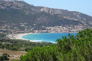 Blick auf eine türkisfarbene Bucht mit Sandstrand, Häusern am Ufer und grünen Bergen im Hintergrund.