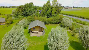 Zelt mit Terrasse auf Holzpodest, Sitzbank und Stühlen, umgeben von Wiesen, Bäumen und einem kleinen Fluss.