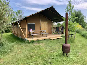 Holzunterkunft mit Terrasse, Bank und Tisch, Markise und Sitzgruppe unter Bäumen auf dem Campingplatz.