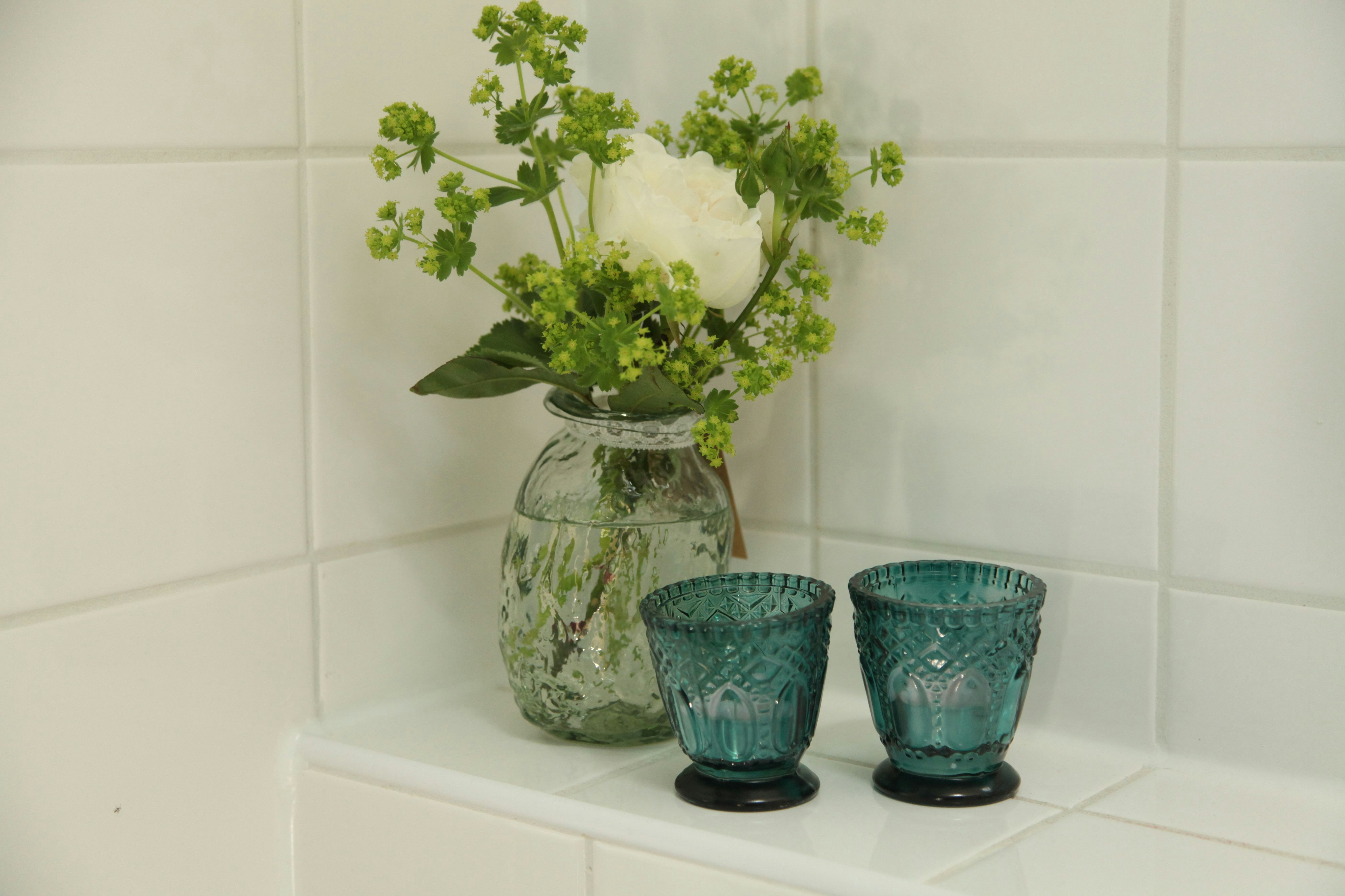 Glasvase mit weißen Blüten und grünen Stielen auf einer weißen Fliesenbank, daneben zwei blaue Teelichtgläser.