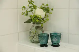 Glasvase mit weißen Blüten und grünen Stielen auf einer weißen Fliesenbank, daneben zwei blaue Teelichtgläser.