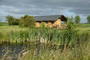 Campingzelt mit Überdachung am Seeufer, Schilf im Vordergrund, grünes Gelände und Gebäude im Hintergrund.