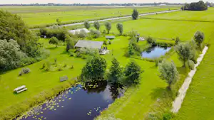 Campingplatz auf grüner Wiese mit Teich, kleinen Gebäuden und Wegen, umgeben von Hecken und Feldern.