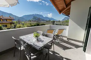 Terrasse mit Tisch und Stühlen, Sonnenschirm, Blick ins Tal und auf Berge, im Hintergrund Häuser und Garten.