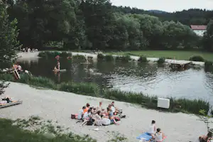 Seeufer mit Kiesstrand, Menschen am Wasser und auf Decken, Ruder am Steg und Bäume im Hintergrund.