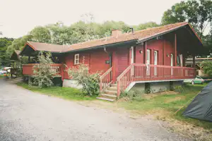 Rotes Holzhaus mit Veranda, Treppe und Dachrinne, daneben Zelt und Auto auf dem Campingplatzweg.