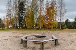 Feuerstelle aus Holz mit Metallring auf einem Campingplatz, umgeben von Wiese und hohen Bäumen im Hintergrund.