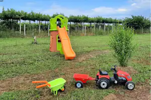 Grüner Rasen mit orange-grüner Kinderrutsche, daneben ein rotes Spielzeugtraktor und eine grüne Schubkarre.