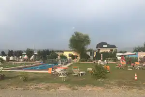 Campingplatz mit Swimmingpool, Liegen und Sonnenschirmen im Garten, daneben Wege, Bäume und ein Gebäude im Hintergrund.
