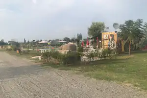 Wohnwagen mit kleiner Terrasse und Sitzgelegenheiten, umgeben von Grün und Hecken; Kiesweg und weitere Unterkünfte im Hintergrund.
