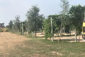 Campingplatz mit Rasenfläche, Baumreihe und Wegen, im Hintergrund Gebäude und ein Wohnwagen am Rand.