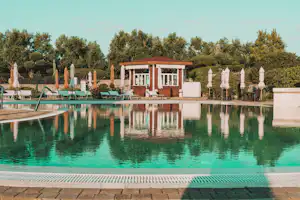 Schwimmbad mit türkisfarbenem Wasser, Liegen und Sonnenschirmen am Beckenrand, dahinter ein Gebäude und Bäume.
