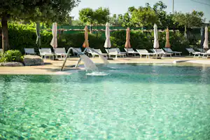 Schwimmbecken eines Campingplatzes mit Liegen und Sonnenschirmen am Beckenrand, im Hintergrund Bäume und Hecken.