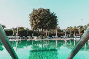 Schwimmbecken mit türkisfarbenem Wasser, Liegestühlen am Rand und Bäumen im Hintergrund.