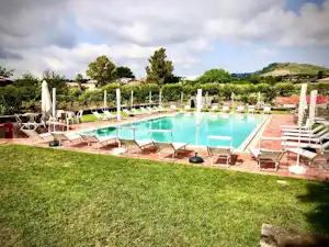 Schwimmbad mit mehreren Liegestühlen und Sonnenschirmen auf einer Rasenfläche, im Hintergrund Bäume und Hügel.