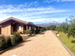 Reihen von Holzbungalows am Campingplatz mit gepflastertem Weg, Büschen und Blick auf Berge in der Ferne.