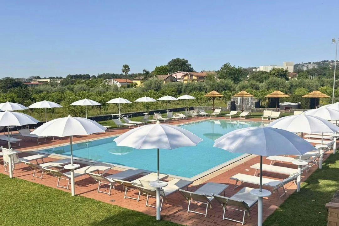 Poolbereich mit türkisfarbenem Wasser, Sonnenliegen und Sonnenschirmen auf einer Rasenfläche, im Hintergrund Gebäude und Bäume.