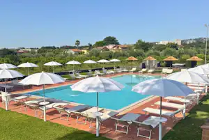 Poolbereich mit türkisfarbenem Wasser, Sonnenliegen und Sonnenschirmen auf einer Rasenfläche, im Hintergrund Gebäude und Bäume.