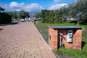 Ziegel-Wasseranschluss mit Zapfhahn am Rand eines gepflasterten Wegs, daneben Rasen und Bäume vor dem Campingplatz.