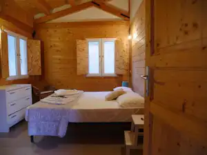 Schlafzimmer in einer Holzunterkunft mit Doppelbett, Nachttisch, Kommode und großen Fenstern, Blick in den Raum.