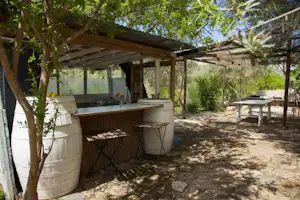 Schattenreiche Terrasse mit Grill, Holzbänken und Tischen, umgeben von Bäumen und Pflanzen im Campingbereich.