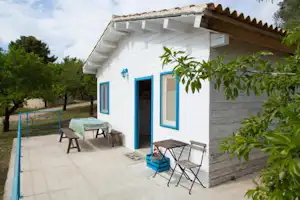 Weiße Ferienhaus-Terrasse mit Tisch und Stühlen, blauen Fenstern und Eingangstür, daneben Pflanzen und gepflasterter Weg.