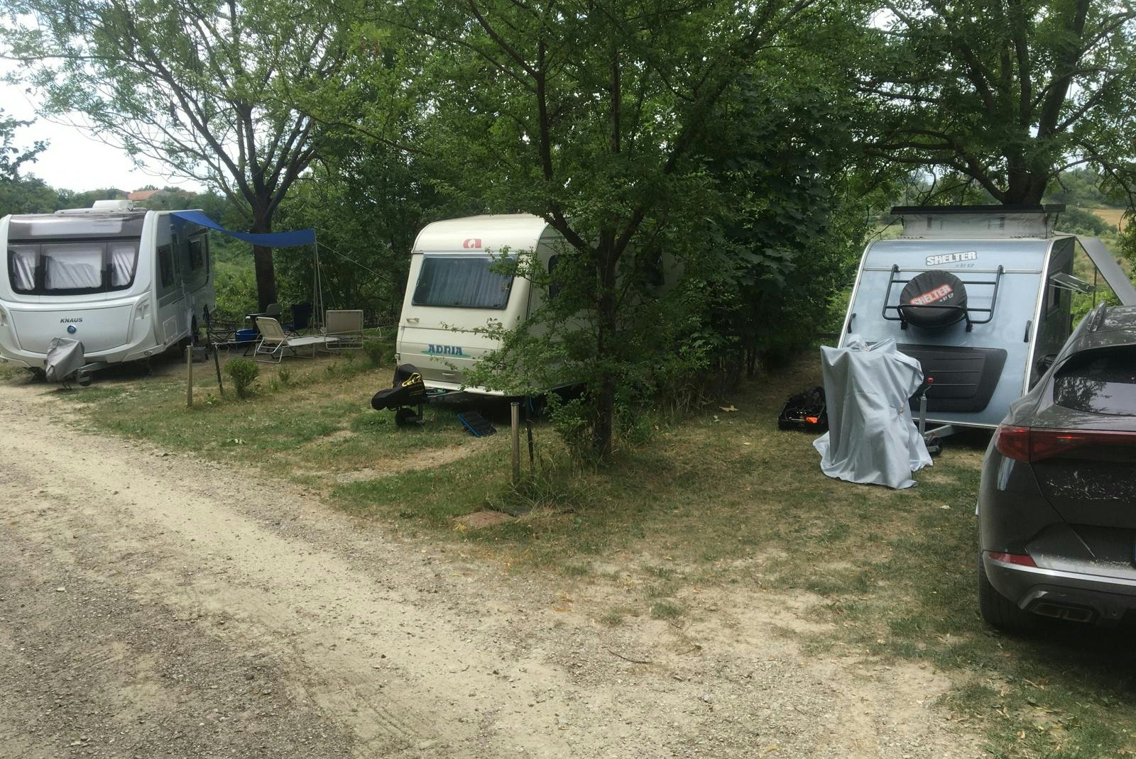 Wohnwagen auf einem Campingplatz-Stellplatz im Grünen, mit Bäumen, Schotterweg und einem Auto am Rand.
