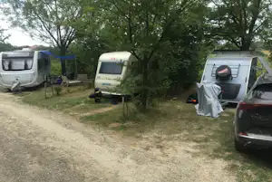 Wohnwagen auf einem Campingplatz-Stellplatz im Grünen, mit Bäumen, Schotterweg und einem Auto am Rand.