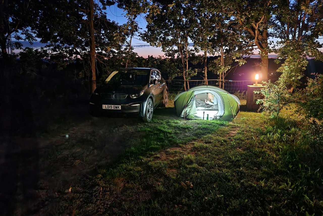 Campingzelt im Wald neben einem Auto auf dem Stellplatz, abends mit Licht am Zelt und Bäumen im Hintergrund.