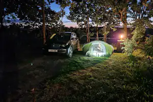 Campingzelt im Wald neben einem Auto auf dem Stellplatz, abends mit Licht am Zelt und Bäumen im Hintergrund.