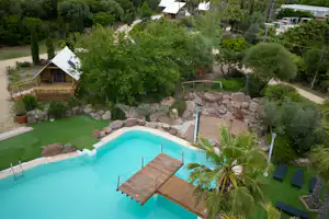 Blick auf den Camping-Pool mit Holzliegen, Palmen und Steinmauer, umgeben von gepflegtem Garten und Wegen.