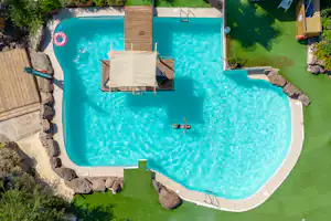 Türkisfarbenes Schwimmbecken auf einem Campingplatz, mit Liegen auf einer Holzplattform und Rasenfläche rundherum.