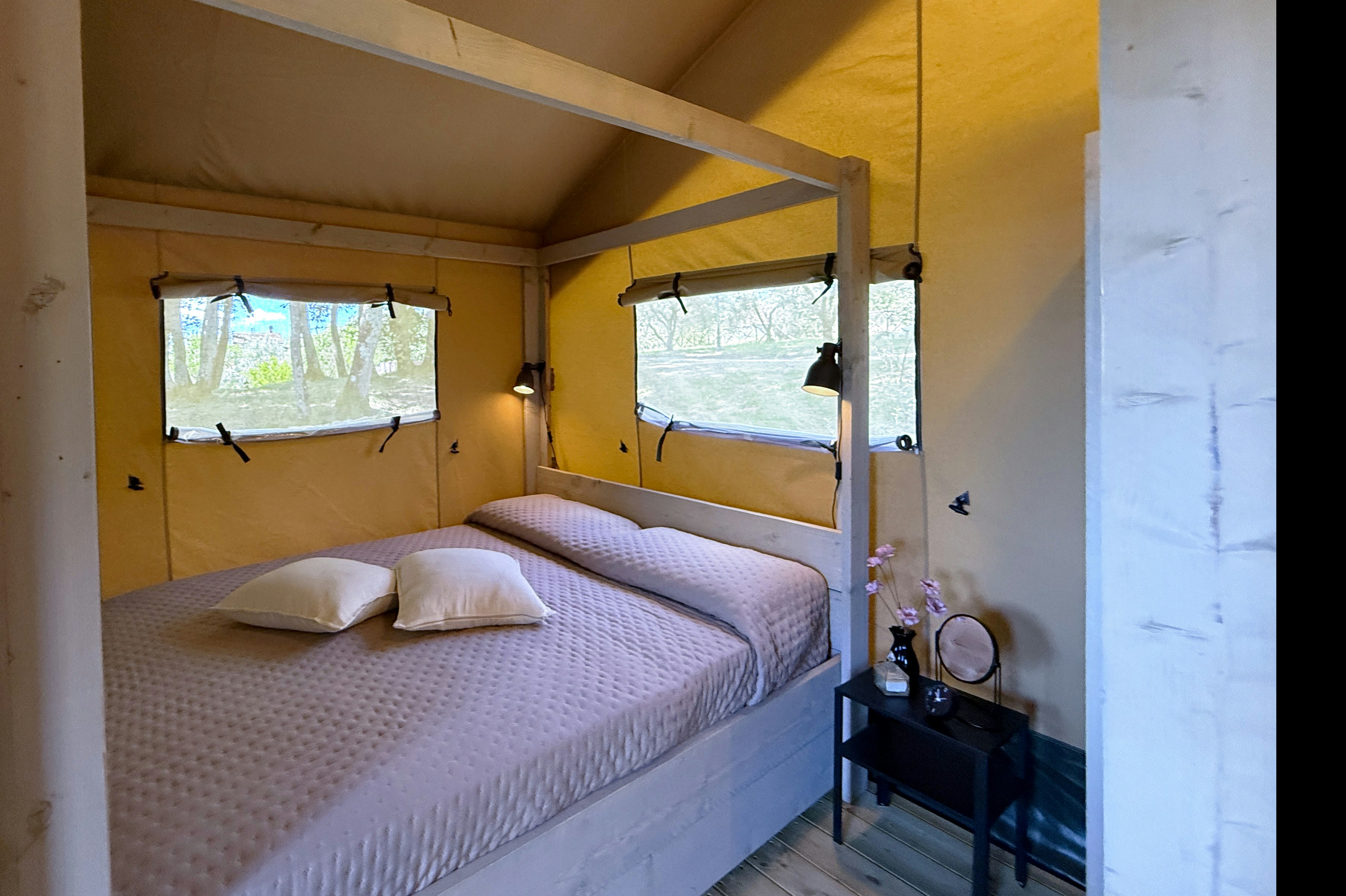 Innenansicht eines gelben Zelts mit Doppelbett, Fenstern mit Vorhängen und Nachttisch mit Lampe.