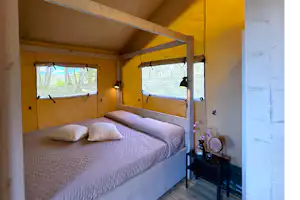 Innenansicht eines gelben Zelts mit Doppelbett, Fenstern mit Vorhängen und Nachttisch mit Lampe.
