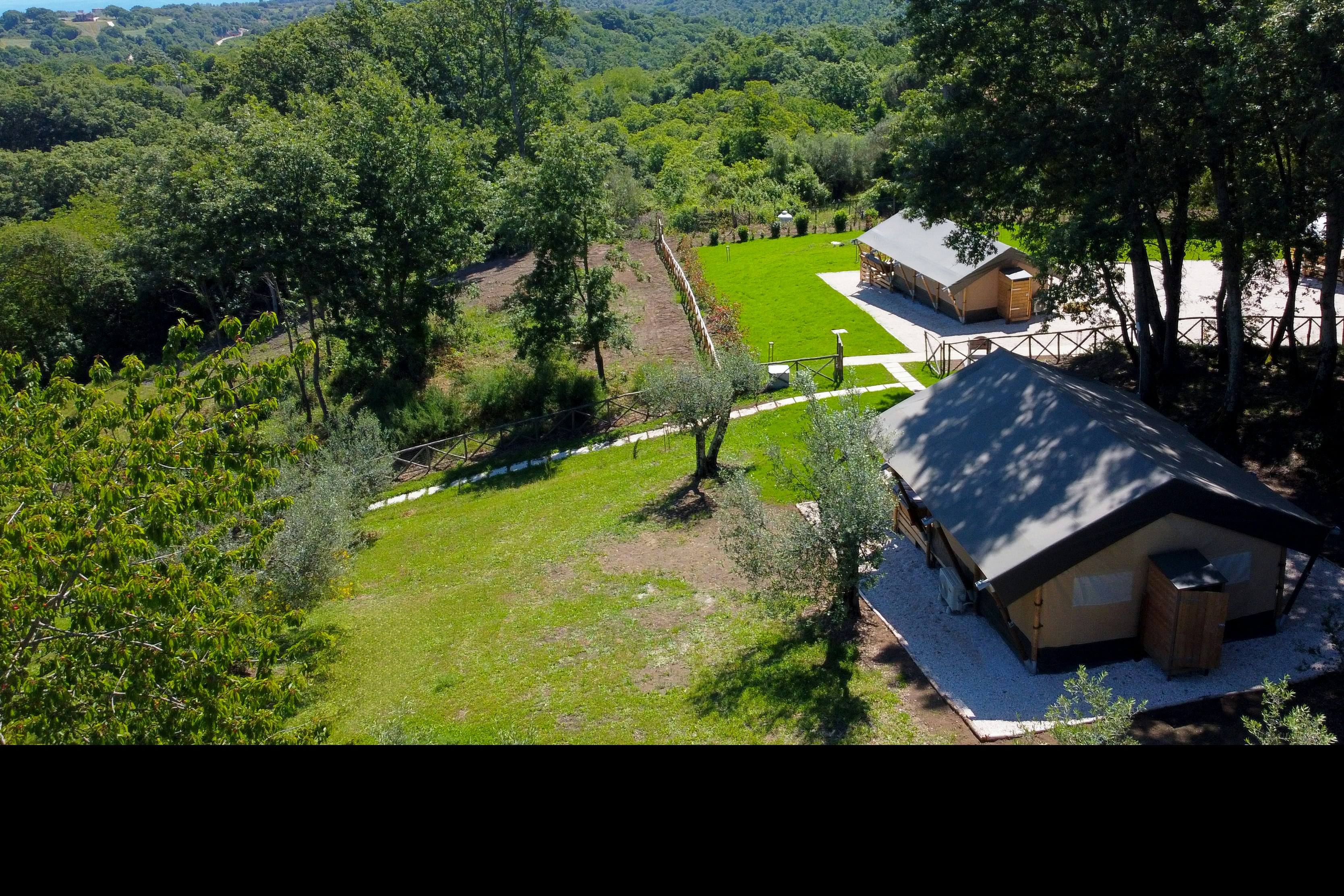 Blick von oben auf mehrere Camping-Unterkünfte mit Holzfassaden, umgeben von Wiesen, Wegen und Bäumen, mit weitem Tal im Hintergrund.