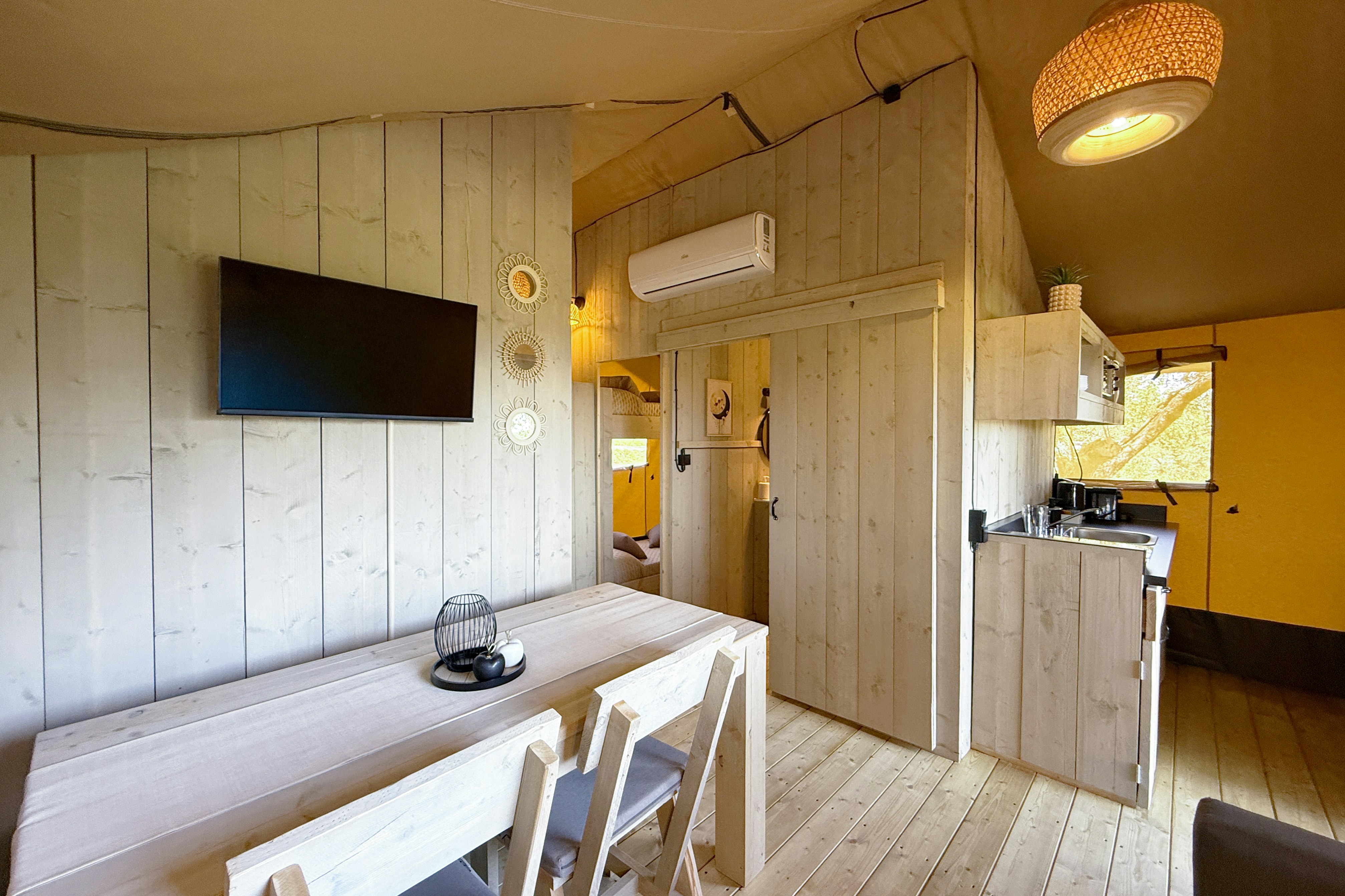 Innenansicht eines Wohnwagens mit Küchenzeile, Esstisch und weißen Stühlen, Holzverkleidung und Flachbildschirm an der Wand.