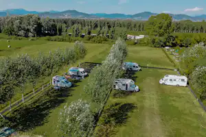 Wohnwagen und Campingfahrzeuge auf einer grünen Wiese, umgeben von Bäumen, mit Blick auf Berge in der Ferne.