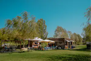 Campingplatz mit mehreren Mobilheimen und Vorzelten, Sitzgruppen und Tischen auf dem Rasen, umgeben von Bäumen.