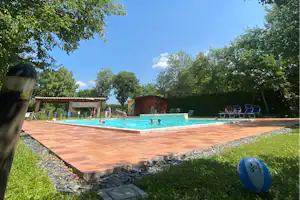 Camping-Pool mit türkisfarbenem Wasser, Terrassenboden und Liegen im Grünen, umgeben von Bäumen und kleinen Gebäuden.