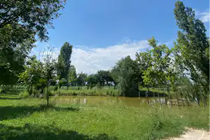 Ruhiger Campingsee mit grünem Ufer, Bäumen und Zaunpfosten im Vordergrund, dazu blauer Himmel mit Wolken.
