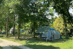 Campingzelt mit Tisch und Stühlen auf dem Stellplatz, daneben Wohnmobil und weitere Zelte zwischen hohen Bäumen.