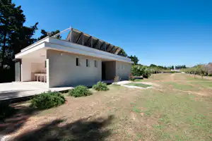 Modernes Campinghaus mit Terrasse, Rasenfläche und Heckenbeeten, daneben ein Holzsteg und Bäume am Rand des Platzes.