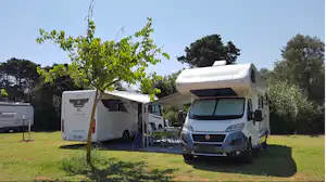 Zwei Wohnmobile auf einem Campingplatz mit Vorzelt, Tisch und Stühlen im Grünen, umgeben von Bäumen.