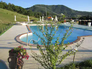 Camping-Pool mit Liegen und Sonnenschirmen, umgeben von Wegen, Pflanzen und Blick auf grüne Berge.