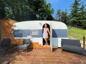 Wohnwagen mit offener Tür auf einer Holzterrasse, zwei Liegesessel davor, umgeben von Bäumen im Grünen.